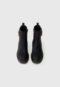 Bottines en cuir noir avec une finition texturée, zip sur le côté et accents en matériel doré. Semelle en caoutchouc noir unie et languette à l'arrière.