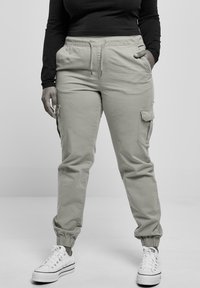 Pantalon cargo gris clair avec une ceinture élastique, des poches latérales et des poignets resserrés, associé à des baskets blanches. Texture de tissu lisse.