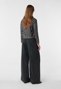 Femme aux longs cheveux bruns portant une veste noire pailletée et un pantalon gris foncé à jambes larges, se tenant de dos contre un fond uni.