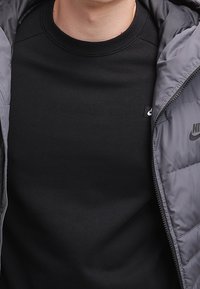 Svart t-shirt med rund hals som bärs under en grå puffjacka. T-shirten har en liten logotyp på framsidan. Jackan har en slät yta.