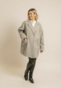 Manteau gris à double boutonnage en tissu texturé, avec de grandes poches et des boutons dorés, associé à un pantalon en cuir noir et des bottes.