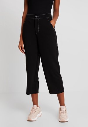 Frau trägt hochtaillierte schwarze, verkürzte Hosen mit weitem Bein und weißen Nähten, kombiniert mit hellrosa klobigen Sneakern und einem schwarzen Oberteil.