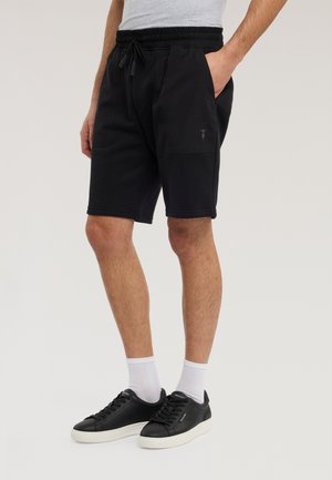 Mężczyzna w czarnych szortach z troczkami, białych skarpetkach i czarnych tenisówkach z białą podeszwą, stojący z jedną ręką w kieszeni.