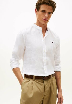 Mężczyzna w białej koszuli zapinanej na guziki z podwiniętymi rękawami oraz beżowych plisowanych spodniach z brązowym paskiem, stojący na tle jednolitej białej ściany.