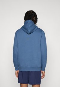 Blauer Kapuzenpullover aus weichem Stoff; verfügt über eine Kängurutasche, gerippte Bündchen und Saum. Die Rückansicht zeigt die Kapuze und die glatte Textur.