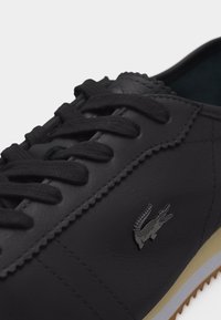 Μαύρο δερμάτινο αθλητικό παπούτσι με μαύρα κορδόνια, ορατή ραφή, μικρό λογότυπο κροκοδείλου στο πλάι και λαστιχένια σόλα σε μπεζ και λευκό χρώμα.