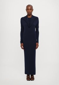 Dlouhé rukávy, tmavě modré šaty s přehozeným výstřihem, slim fit a dlouhým střihem až na zem, doplněné o sandály na volné šněrování v černé barvě. Hladká textura.