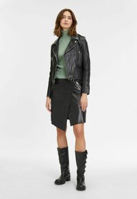 Schwarze Lederjacke mit silbernen Beschlägen, grüner gerippter Rollkragenpulli, schwarzer Wickelrock aus Kunstleder und knielange schwarze Stiefel mit Schnallen.