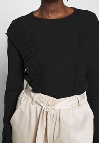 Haut noir à manches longues avec des accents texturés et volantés le long des côtés, associé à un pantalon taille haute de couleur crème avec une ceinture nouée.