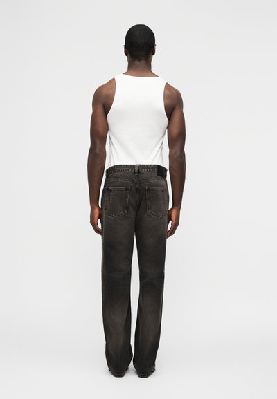 Άνδρας με την πλάτη γυρισμένη, φορώντας λευκό αμάνικο και φαρδιά μαύρα ξεθωριασμένα τζιν, στέκεται μπροστά σε απλό ανοιχτό γκρι φόντο.