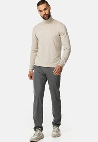 Beżowy sweter z wysokim kołnierzem w połączeniu z szarymi spodniami. Materiał wydaje się miękki i gładki. Model nosi jasne sportowe buty z sznurówkami, stojąc na neutralnym tle.