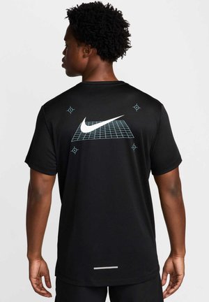 Mand med kort krøllet hår iført sort Nike T-shirt med hvid swoosh over blåt gitter og målsymboler på ryggen, stående mod hvid baggrund.
