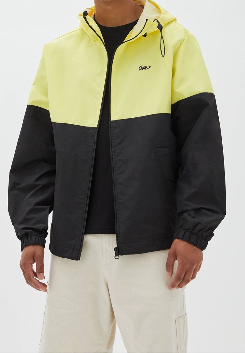 Homme portant une veste coupe-vent jaune et noire avec capuche, chemise noire et pantalon beige clair, debout les bras détendus.