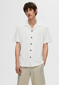 Mann iført en hvit kortermet skjorte med knapper og beige shorts, stående med den ene hånden i lommen mot en ensfarget bakgrunn.