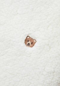 Vloeiend wit materiaal met een klein, geborduurd bruin berenhoofd. De textuur oogt zacht en pluizig, wat visuele interesse toevoegt.
