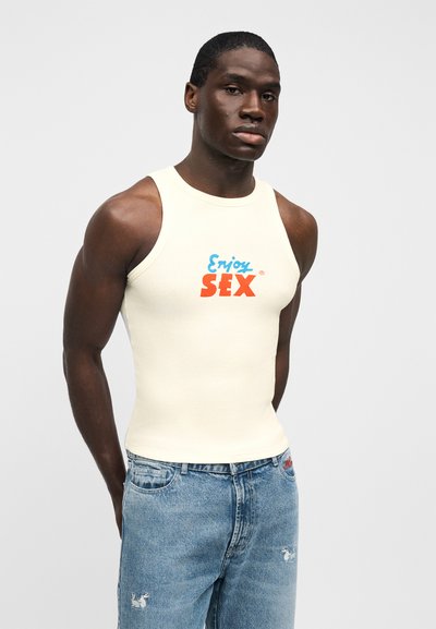 Κρεμ ριμπ τοπ με μπλε και κόκκινο γραφικό κείμενο «Απόλαυσε το ΣΕΞ», συνδυασμένο με ανοιχτό μπλε distressed τζιν σορτς.