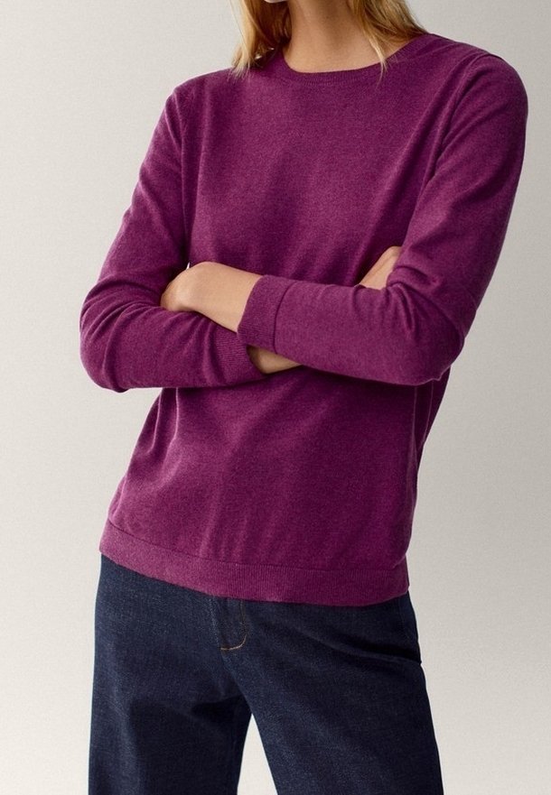 Femme aux bras croisés portant un pull violet à manches longues et un jean bleu foncé sur un fond uni.