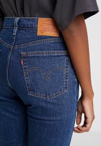 Vaqueros de mezclilla azul de cintura alta, con un parche de cuero en la parte trasera, costuras en contraste y un diseño distintivo en el bolsillo trasero.