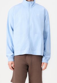 Ljusblå zip-up jacka med ståkrage, två framfickor, elastiska manschetter och en strukturerad tygkvalitet. Har en liten logotyp på bröstet.