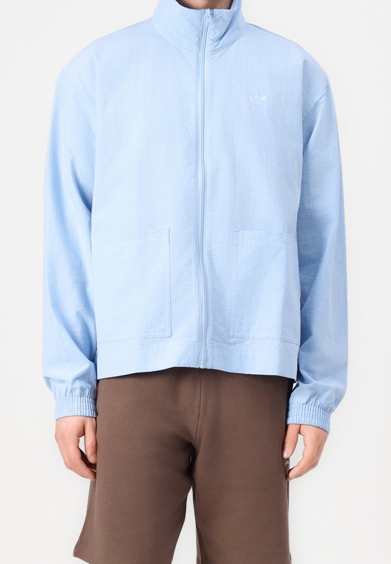 Ljusblå zip-up jacka med ståkrage, två framfickor, elastiska manschetter och en strukturerad tygkvalitet. Har en liten logotyp på bröstet.