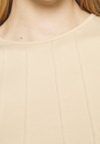 Närbild av en persons nacke och övre bröst som bär en beigh ribbat tyg topp med rund halsringning.