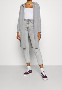 Długi szary kardigan z kapturem, dwoma przednimi kieszeniami, noszony na białym topie na ramiączkach. W zestawieniu z jasnoszarymi dżinsami i bordowymi sneakersami.