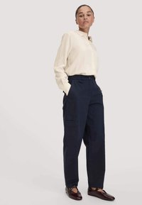 Vrouw met donker haar in een staart, gekleed in een crèmekleurig shirt met lange mouwen, marineblauwe high-waist broek en donkere leren flats, staand met haar handen in de zakken.