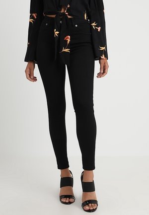 Zwarte skinny jeans met een gladde textuur en een strakke pasvorm, gecombineerd met zwarte block-heeled sandalen en een top met bloemenprint.