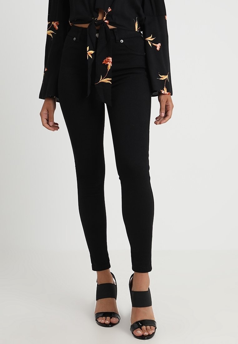 Vaqueros negros ajustados con una textura suave y un diseño ceñido, combinados con sandalias negras de tacón cuadrado y una blusa de estampado floral.