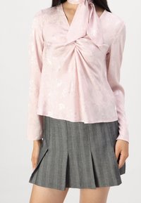 Blusa rosa chiaro con motivo floreale e maniche lunghe, dotata di un dettaglio annodato davanti. Abbinata a una gonna grigia plissettata con righe verticali.