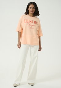 T-shirt oversize pêche avec logo rouge "LEISURE League" imprimé ; associé à un pantalon ample blanc et des chaussures beiges à enfiler.