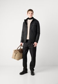 Schwarze, gepolsterte Jacke mit hohem Kragen, cremefarbener Strickpullover, dunkle Hose und schwarze Stiefel. Hält eine beige und braune Sporttasche.