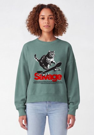 Junge Frau mit lockigem Haar, die einen grünen Pullover mit einer schwarzen Katze auf einem Skateboard und dem Wort "Savage" in rotem Text trägt.