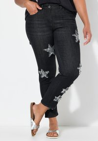 Vaqueros de mezclilla negra con parches de estrellas desgastados y bordes deshilachados. Combinados con sandalias deslizables plateadas. Diseño ajustado y cónico.