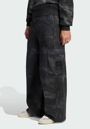 Person trägt lockere schwarze Camo-Cargohose mit Seitentasche und Adidas-Logo, kombiniert mit hellen Sneakern und einem dunkel gemusterten Sweatshirt.