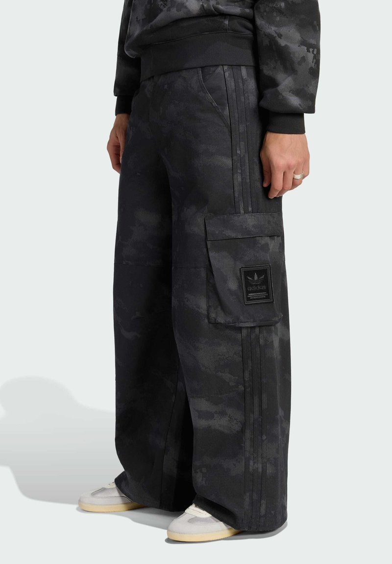 Person wearing loose black camo cargo pants with side pocket and Adidas logo, paired with light sneakers and a dark patterned sweatshirt.