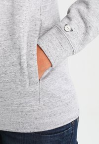 Szary sweatshirt o teksturowanej powierzchni, z kieszenią boczną i ściągaczami na rękawach. Zauważalny akcent w postaci srebrnego okucia na mankiecie.