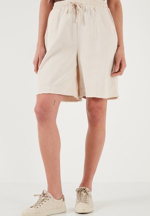 Beige shorts med elastisk talje, snor og frynset kant, båret med beige lave sneakers på en stående person.