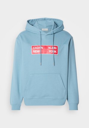 Light blue hoodie with a front pocket, featuring a bold red rectangular graphic with the text "CALVIN KLEIN NEW YORK" in white. Adjustable hood.
