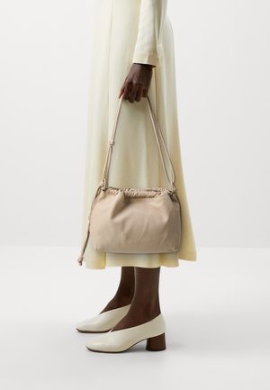 Borsa in tessuto beige e morbido con una parte superiore arricciata, manico annodato e forma arrotondata. Indossata con un vestito giallo chiaro e scarpe con tacco bianche.