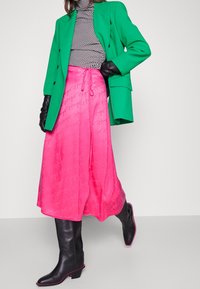 Zelený oversized blazer, černý a bílý vzorovaný rolák, růžová saténová maxi sukně s logem, a černé kozačky po kolena s růžovými akcenty.