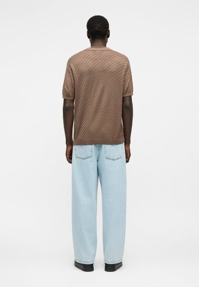 Homme de dos portant une chemise marron à manches courtes texturée, un jean bleu clair à jambes larges et des chaussures noires sur un fond uni.