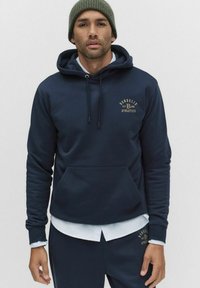 Mężczyzna ubrany w granatowy hoodie i dopasowane dresy z żółtym logo "Bondelid Athletics", nałożone na biały t-shirt i zieloną wełnianą czapkę.