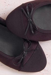 Par mørkelilla ballerinaer med sorte sløjfer på tæerne, med teksturerede og glatte stofsektioner.