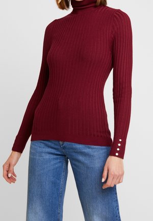Femme portant un pull à col roulé côtelé bordeaux ajusté avec des boutons sur les poignets des manches et un jean bleu en denim.
