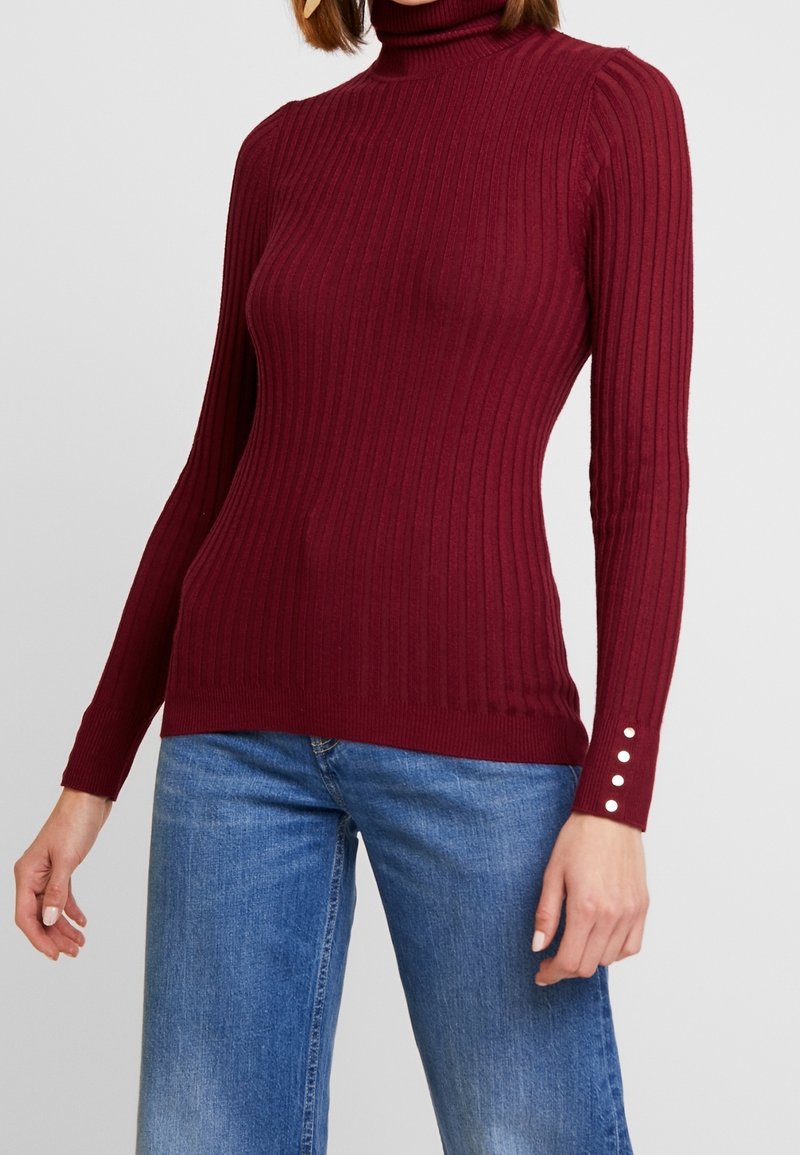 Femme portant un pull à col roulé côtelé bordeaux ajusté avec des boutons sur les poignets des manches et un jean bleu en denim.