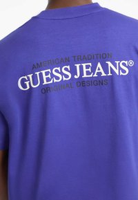 Vijolična bombažna majica s kratkimi rokavi, na hrbtni strani pa so v belih in črnih črkah napisani "AMERIČNA TRADICIJA GUESS JEANS ORIGINALNI ODMETI."