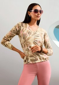 Frau trägt übergroße pinke Sonnenbrille, beige Sweater mit Blumenmuster, pinke Hose und ein braunes Armband, posiert mit einer Hand auf der Hüfte.