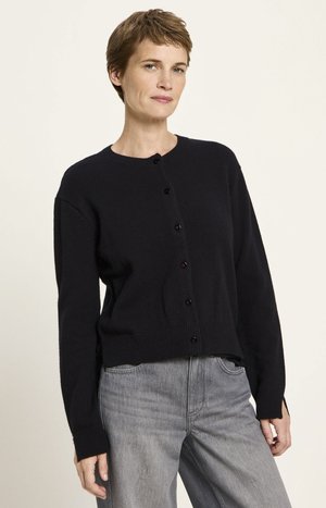 Frau mit kurzen Haaren, die eine schwarze, geknöpfte Strickjacke und graue Jeans trägt, steht vor einem einfarbigen, hellen Hintergrund.