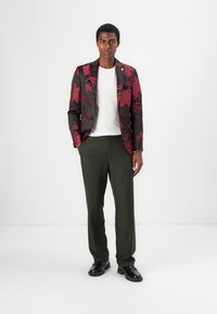 Schwarze und burgunderfarbene Blumenjacke über einem weißen T-Shirt, kombiniert mit grünen Hosen und schwarzen Schuhen. Verfügt über ein tailliertes Design und strukturierte Stoffe.